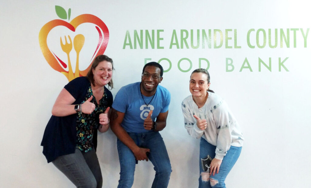 A photo of CTM volunteers Jessica Michaels, James Wicks, and Courtney Tyson in front of an Anne Arundel County Food Bank banner giving a thumbs up.