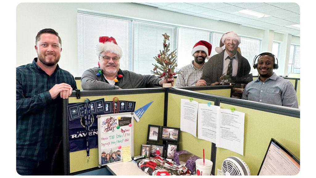 Group of men gather around a cubicle with a cardboard cutout of Clark Griswold showing their collaboration and teamwork. 